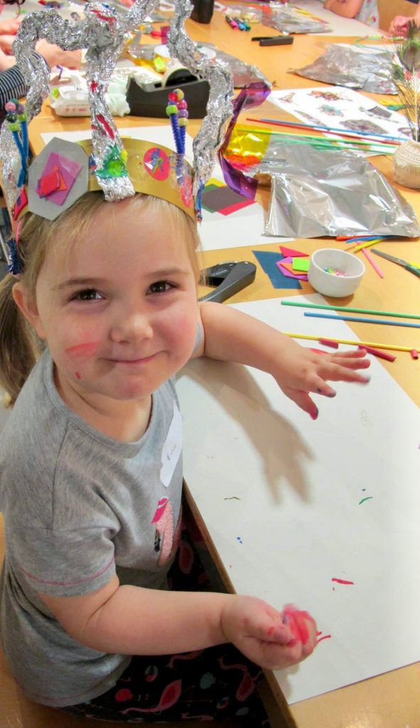 A toddler wearing a crown made from foil and craft materials smiles up at the camera