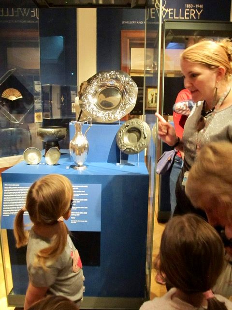 Children explore the exhibition with museum educator Nicola. They look up at a display case containing large silver plates and jugs.
