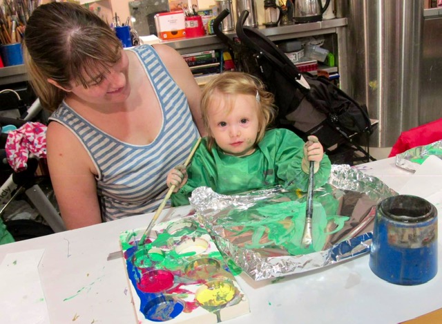 A baby wearing a green overall sits on her mum's lap, painting green paint on foil.