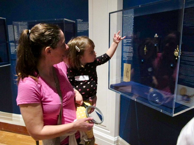 A mum and baby explore a case in the Jewellery exhibition. The case has a dark blue background with gold jewellery displayed inside. 