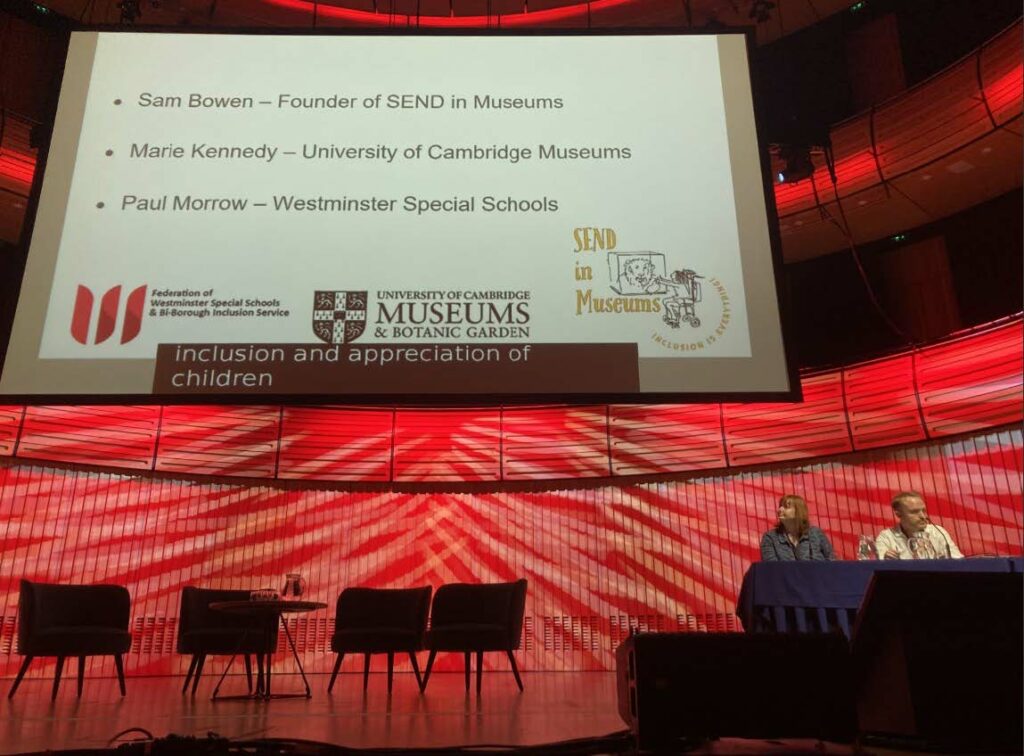 Marie sits next to a man behind a table on stage and woman sitting on stage. Above them is a large projection screen, including their names, job titles and organisation logos.