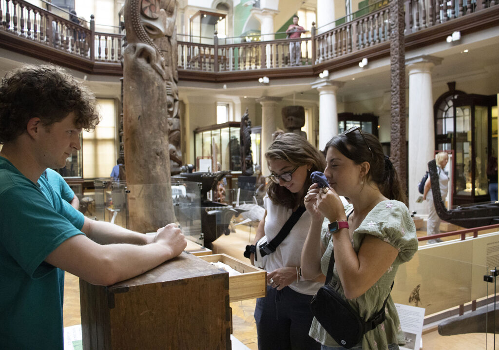 Standing in a gallery at the Museum of Archaeology and Anthropology, a museum intern wearing a blue shirt leans on a wooden box to speak to two visitors who lift up objects and smell them.