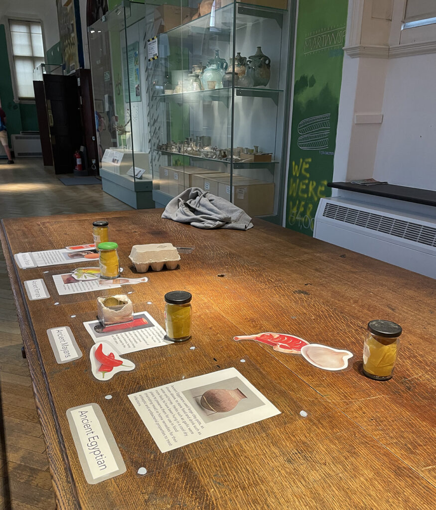 A wooden table in the Andrews Gallery of the Museum of Archaeology and Anthropology covered with A4 signs with information about food in different civilisations, including jars and pictures of different foods.