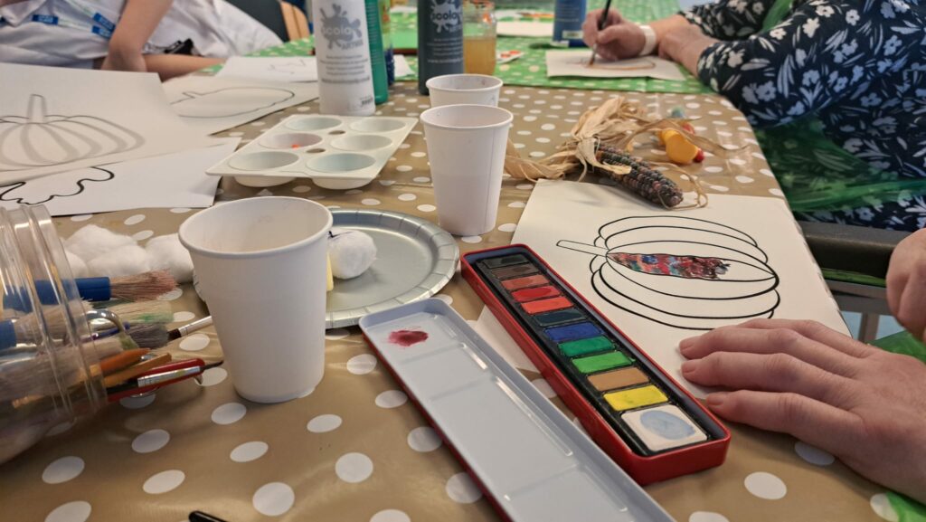 Patients sit around a table covered with a dotty tablecloth and art materials, painting a template of pumpkin.