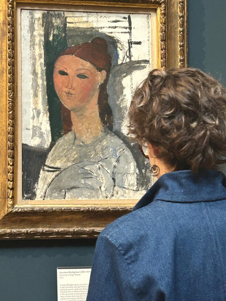 A person with curly brown hair and wearing a blue shirt looks at a gold framed painting of a woman at the Fitzwilliam Museum.