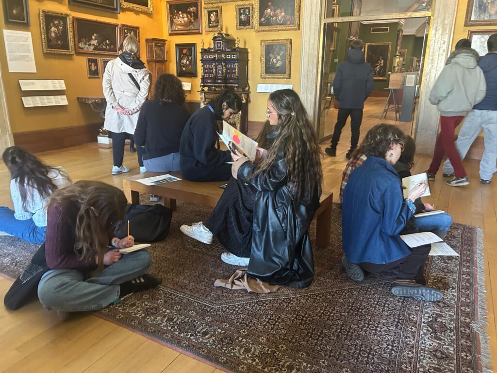 A group of young people sit on a bench and on a rug in the flower paintings gallery at the Fitzwilliam Museum, all drawing or writing in their notebooks.