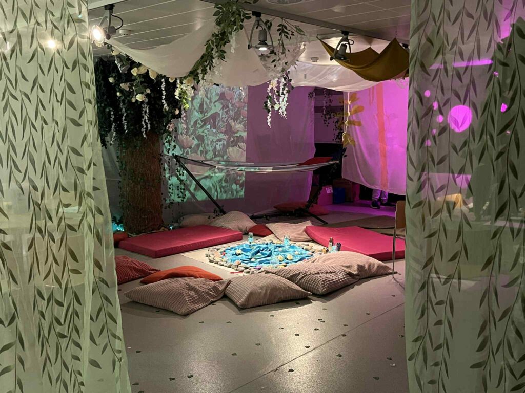 The Happy Place installation with foliage and fabrics hanging from the ceiling and walls, and a circle of soft mats and beanbags in the middle of the floor around a sensory installation. A hammock with a red pillow is in the background. The lighting is soft and coloured.