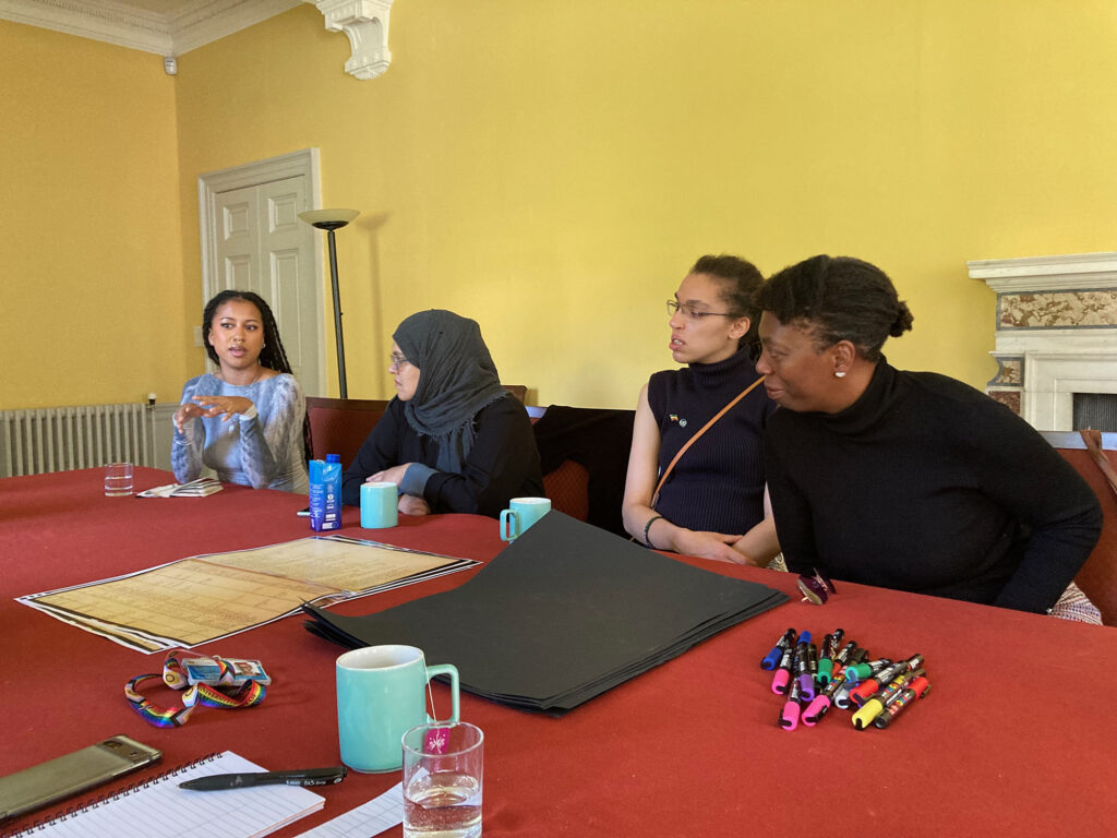 Four people sit talking at a table in the third Black Atlantic Community Group workshop. On the table are papers, pens, mugs and documents.