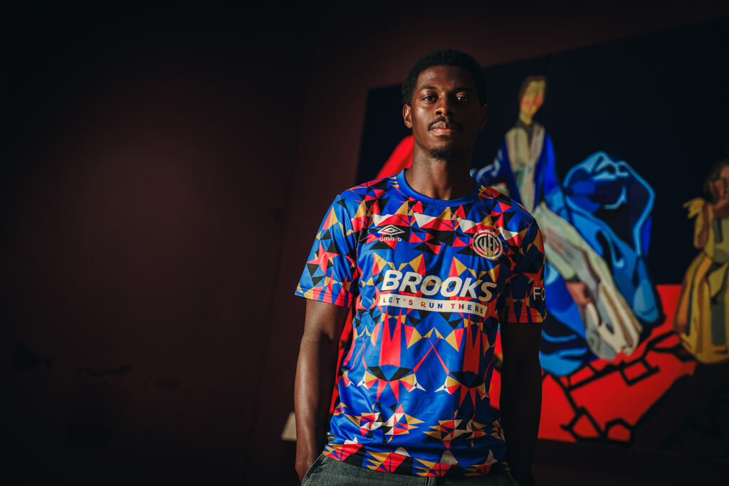 Cambridge United FC player Sullay KaiKai stands wearing the new shirt in front of Joy Labinjo’s An Eighteenth-Century Family which inspired the design.