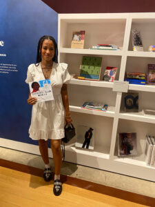 Selena Scott stands smiling in a white dress holding a copy of her colouring book next to a bookcase in the Black Atlantic Reflection Space.