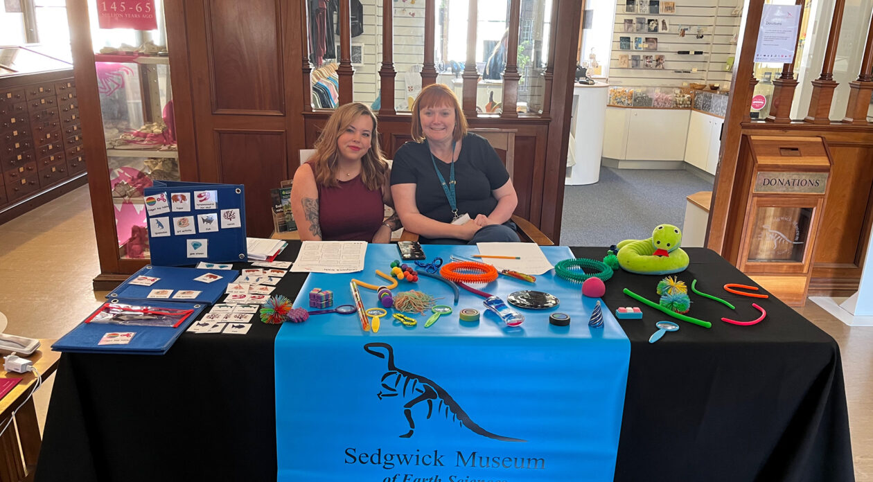 Two women sit in front of a table at the Sedgwick Museum. The table is covered in the museum logo with a dinosaur skeleton and with sensory toys.