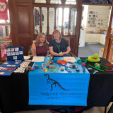 Two women sit in front of a table at the Sedgwick Museum. The table is covered in the museum logo with a dinosaur skeleton and with sensory toys.