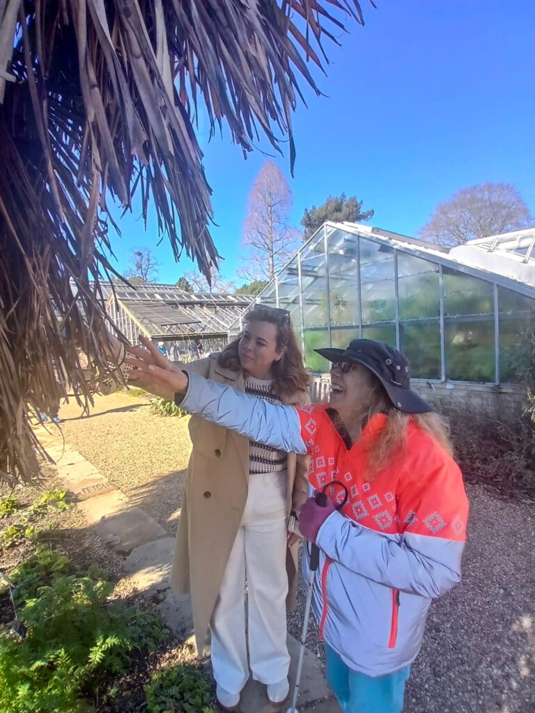 Two women stand together and reach out to feel the leaves of a tree at the Cambridge University Botanic Garden. The glasshouse can be seen in the background.
