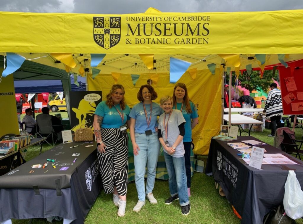 Four women stand smiling inside a bright yellow gazebo with University of Cambridge Museums logo on the front of it.