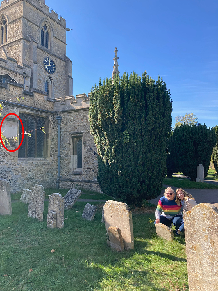 Two women kneel smiling by the footstone next to St Andrew Parish Church on a sunny day. Circled on the left is the engraved epitaph on the church wall.