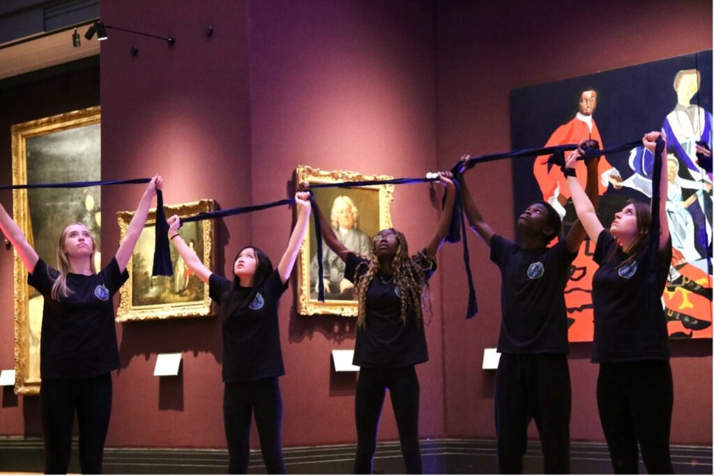 Five young performers stand stretching a piece of dark fabric over their heads during a performance at the Fitzwilliam Museum.