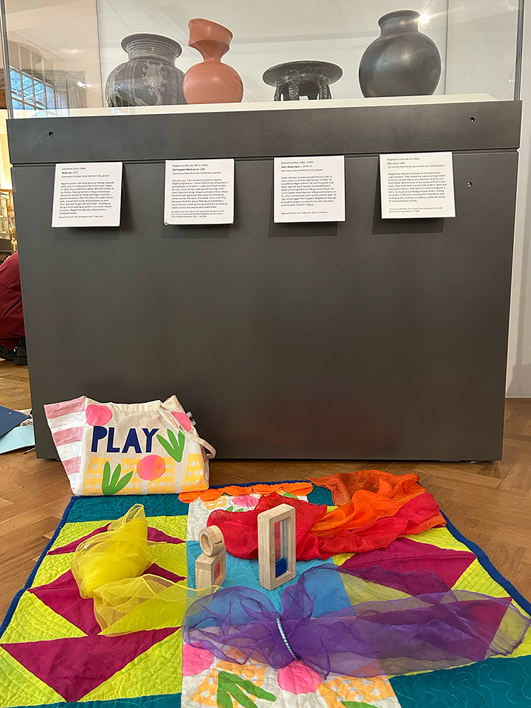 A colourful patterned baby mat spread on the floor in front of a glass display cabinet of pottery at the Fitzwilliam Museum.