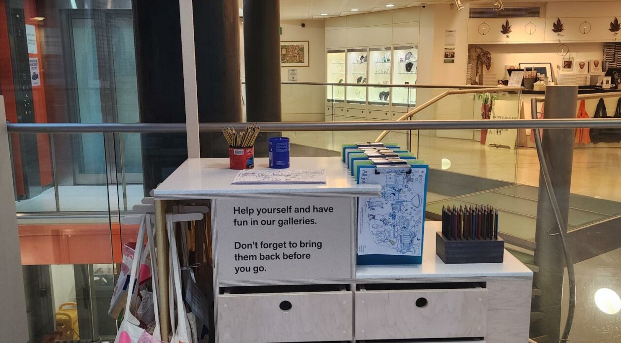 A trolley with drawer on wheels. A lollipop style sign on top of the trolley reads 'Families' and full tote bags hang from one side. In the background is the Fitzwilliam Museum shop and lift.