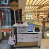 A trolley with drawer on wheels. A lollipop style sign on top of the trolley reads 'Families' and full tote bags hang from one side. In the background is the Fitzwilliam Museum shop and lift.