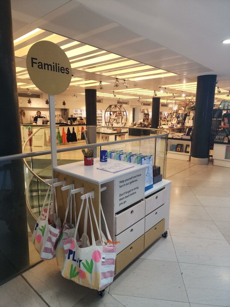 A trolley with drawer on wheels. A lollipop style sign on top of the trolley reads 'Families' and full tote bags hang from one side. In the background is the Fitzwilliam Museum shop.