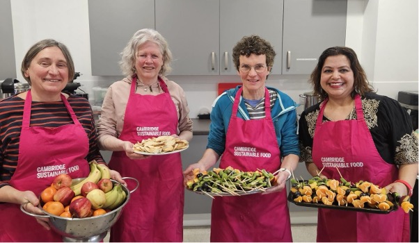 Four people standing holding platters of food and wearing pink Cambridge Sustainable Food aprons.