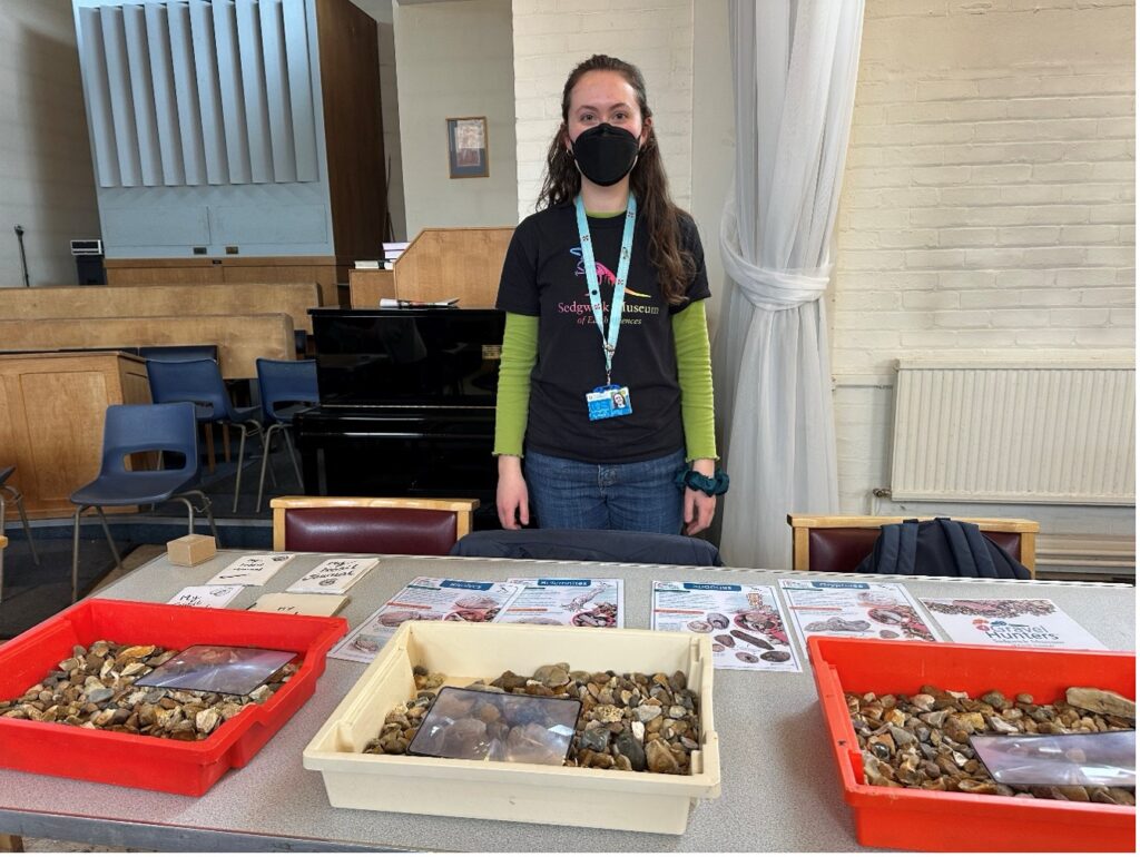A young woman stands in front of a table fulled with fossils and activity sheets at a community lunch event.