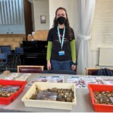 A young woman stands in front of a table fulled with fossils and activity sheets at a community lunch event.