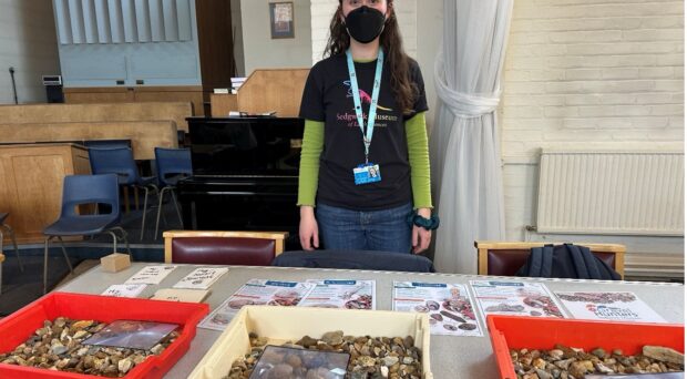A young woman stands in front of a table fulled with fossils and activity sheets at a community lunch event.