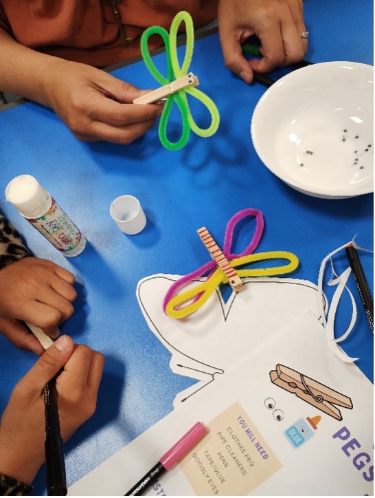 A close up of children taking part in a craft activity, creating pipe cleaner butterfly pegs at a blue table.