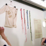 A close up of the Young Producers display with wall-mounted long-sleeved top and weavings, and a sign reading The Gifts with further information. Three young producers' heads are visible looking towards the display.