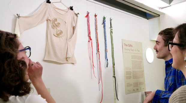 A close up of the Young Producers display with wall-mounted long-sleeved top and weavings, and a sign reading The Gifts with further information. Three young producers' heads are visible looking towards the display.