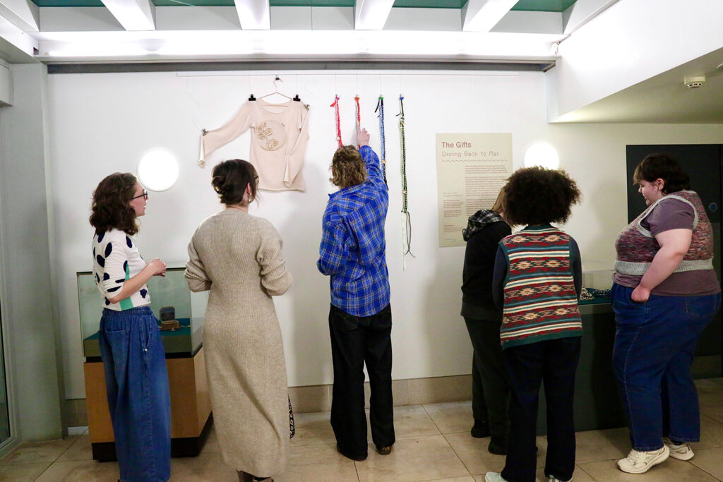 A group of six people stand looking at their display in the Fitzwilliam Museum. A sign on the wall reads The Gifts.