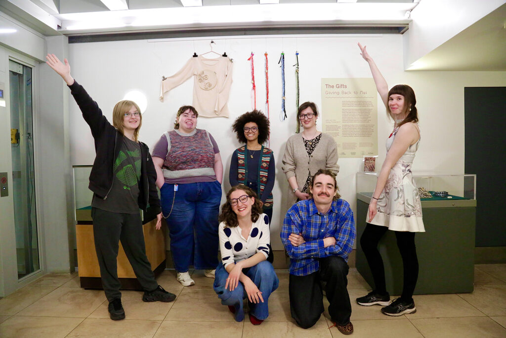 A group of seven smiling young producers, five standing (two with arms in the air) and two kneeling, in front of their display at the Fitzwilliam Museum.