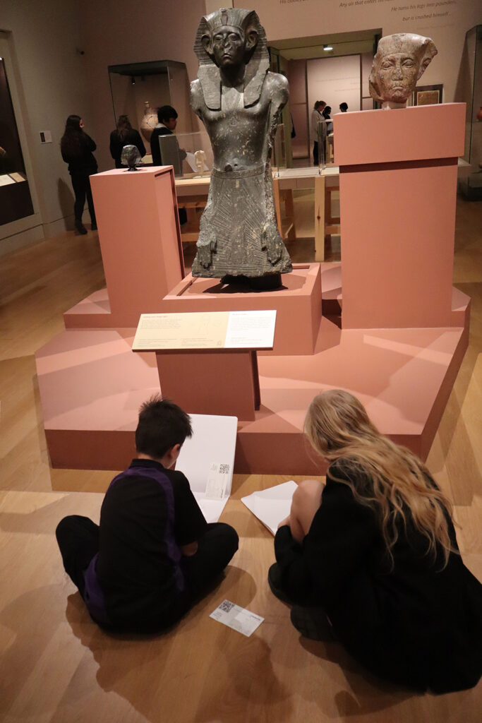 Students sitting on the floor in the ‘Made in Ancient Egypt’ exhibition drawing and facing a statue.