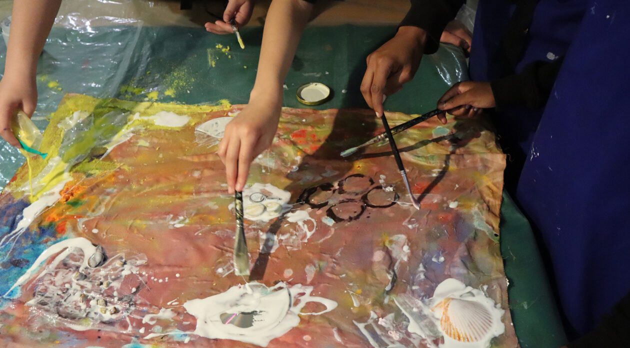 Children's arms holding paintbrushes and working collaboratively on bright abstract pour paintings inspired by Frank Bowling’s abstract paintings.