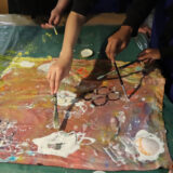 Children's arms holding paintbrushes and working collaboratively on bright abstract pour paintings inspired by Frank Bowling’s abstract paintings.