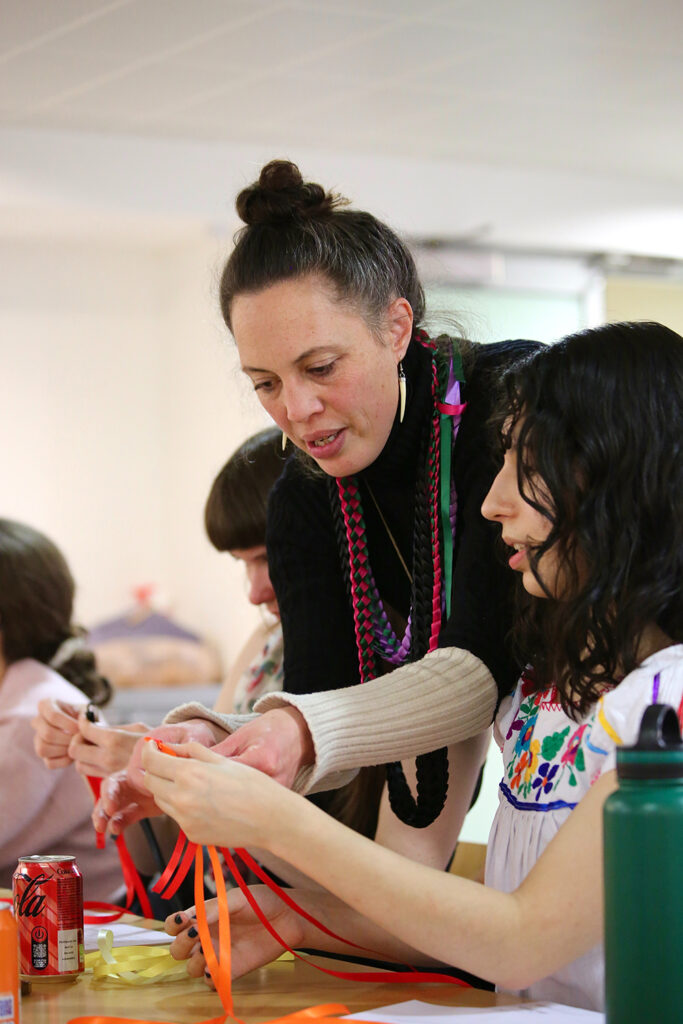 A woman wearing long necklaces bends forward to help a young person at a weaving workshop.