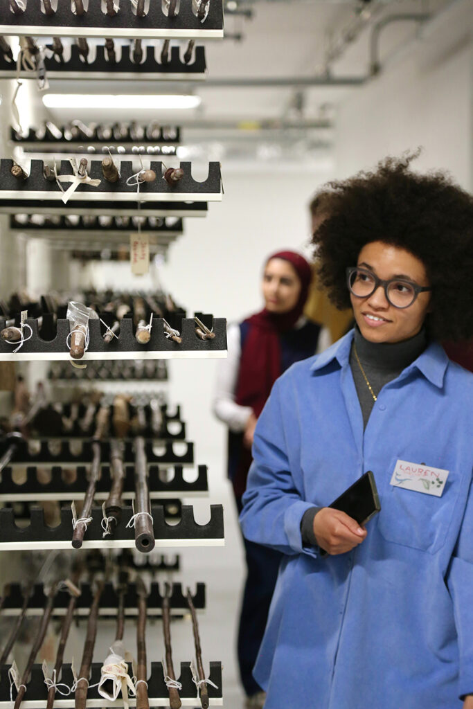 Two young producers, one with an afro and glasses, and another in the background with a red headscarf, next to a display of spears at the Centre for Material Culture.