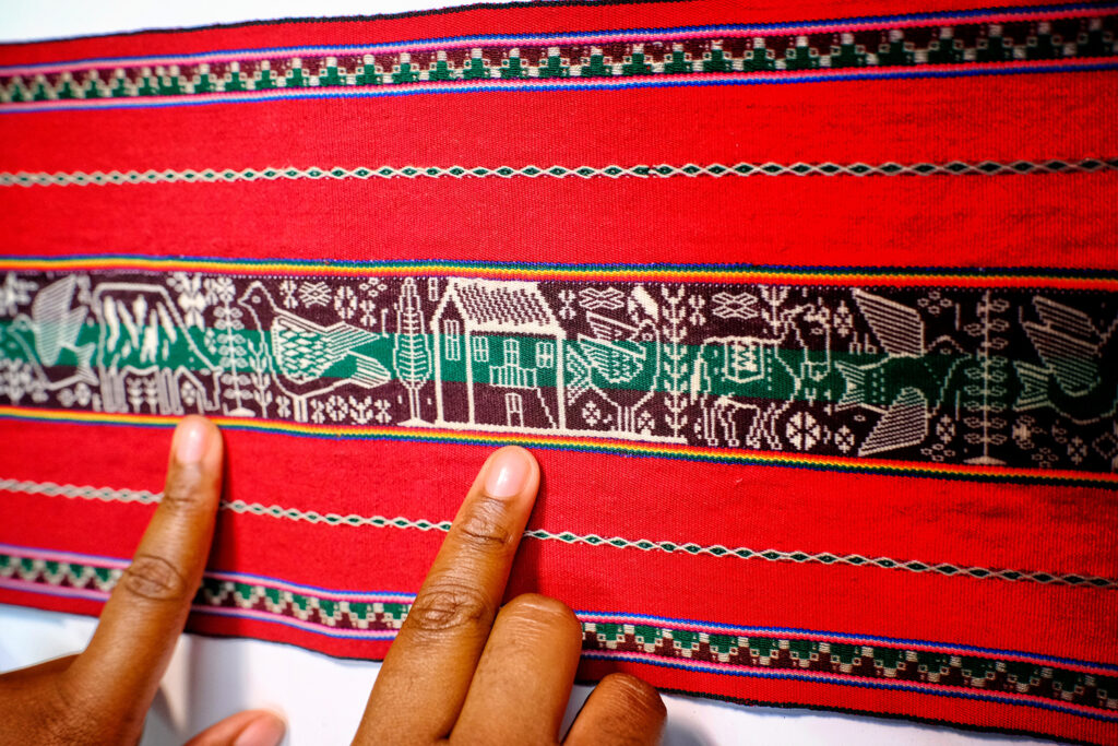 A close up of someone holding both index fingers against a red piece of fabric embroidered with patterned borders and a central band of embroidered designs including bird, horse, a house and trees.