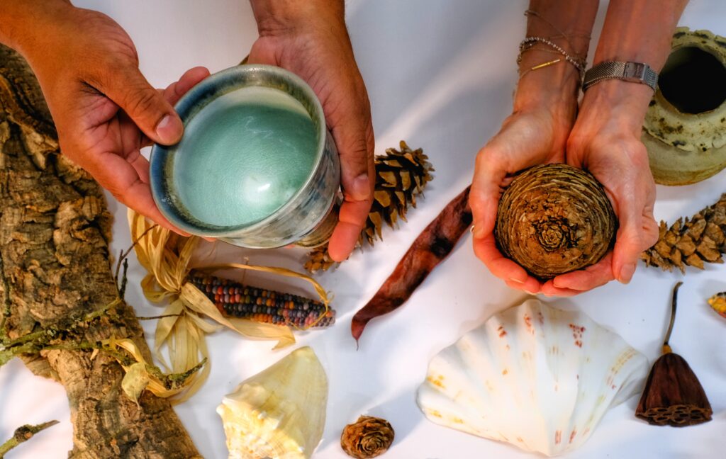 A close up of two pairs hands from above, one holding a ceramic pot and the other a pine cone. There are an array of objects on the white clothed table beneath them.