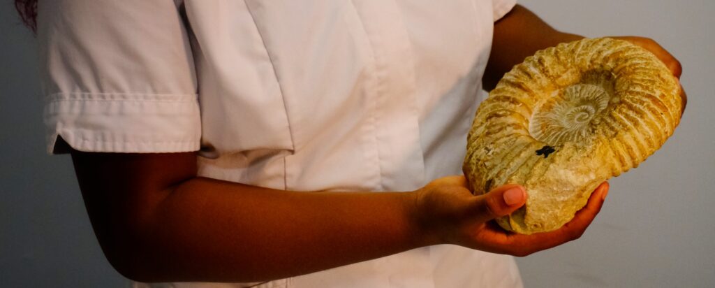A close up of a woman wearing medical clothing holds a large ammonite in both hands.