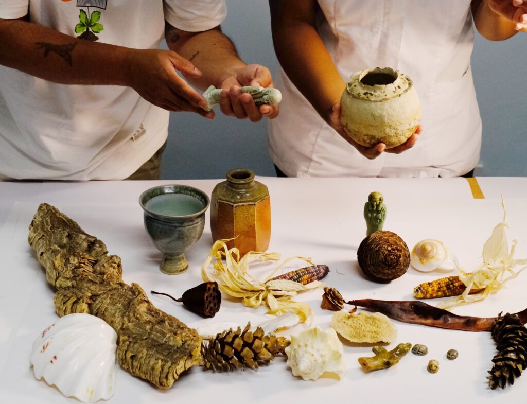 Two people stand in front of a table that holds a variety of objects, including a shell, pots, pine cones and other natural objects. One holds a small figure sculpture, the other a pot.
