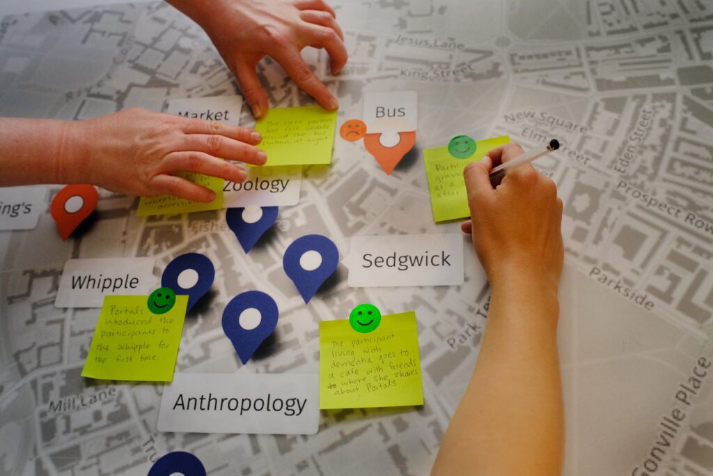 Seen from above, the hands of Katie and a Portals participant be seen placing and writing on post it notes on a black and white map of Cambridge.
