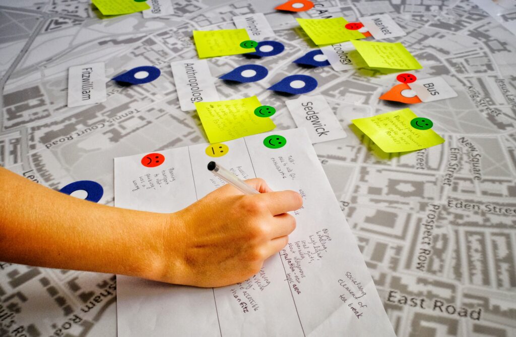 Katie writes notes on a piece of paper labelled with three faces: happy, neutral and sad. Beneath this piece of paper is a black and white map of Cambridge, covered with markers showing the museums and posit notes of participants' feedback.