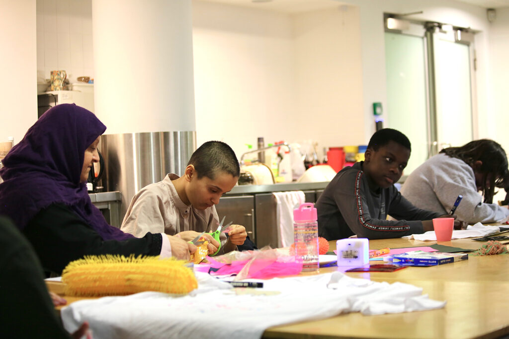 Three young Arts Pioneers participants sit along one side of a wooden table working on crafts at the Fitzwilliam Museum, with a parent sat to their left wearing a purple headscarf. On the table are craft materials: t shirt, sensory toys, drawing materials and a water bottle.
