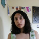 Mia González Janes, a young woman with brown shoulder length wavy hair, looking at the camera. She is say in a room that is decorated with a string of photos and a canvas.