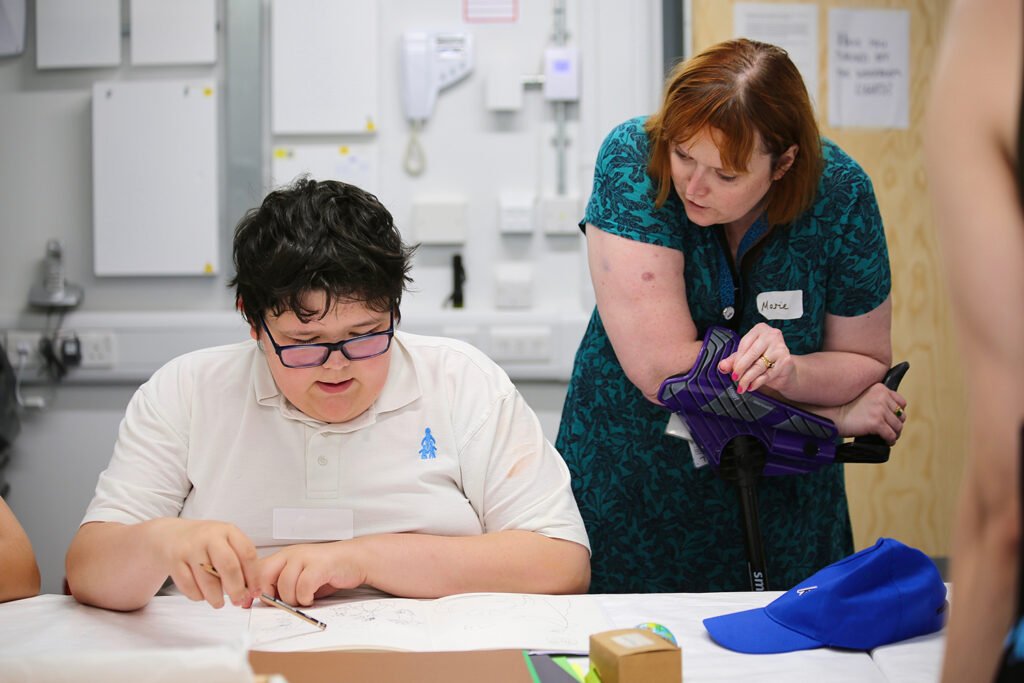 Marie, a woman with short red hair wearing a green dress, leans on her walking stick to talk to a school pupil sat at a desk looking at some work on a table in front of him and pointing at it with a pencil.