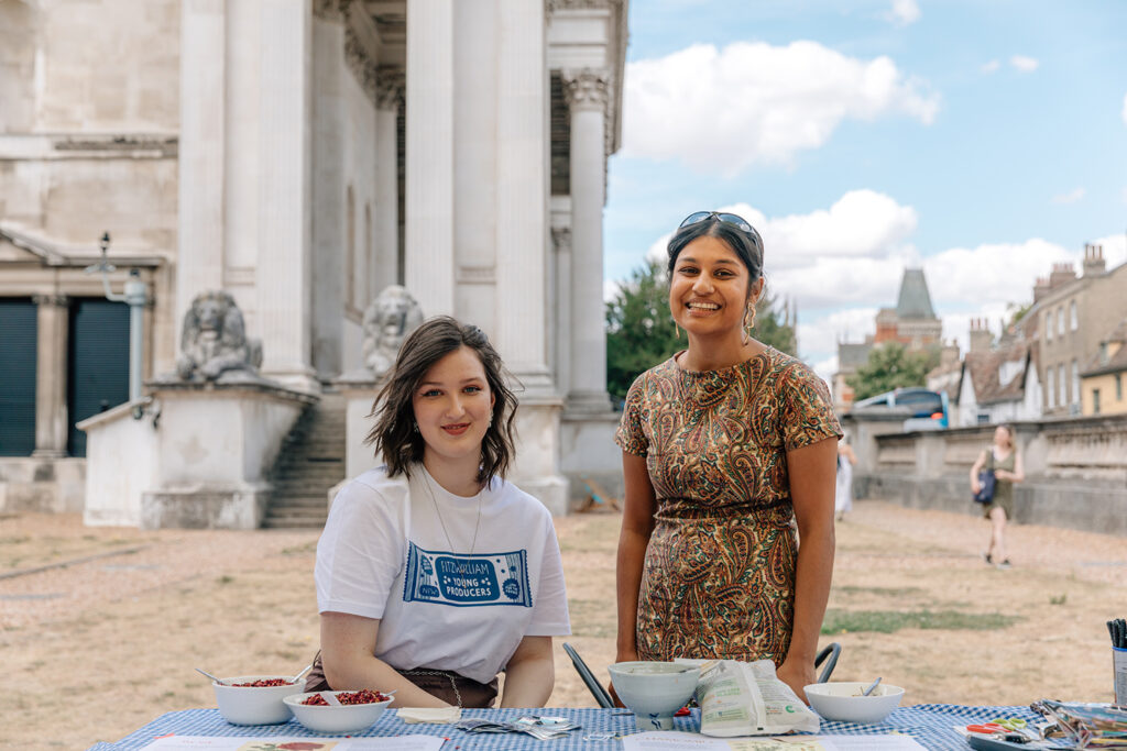 Two women, one sitting and one standing in front of a table, smile on the green outside the Fitzwilliam Museum. In the background are the museum entrance steps, flagged by lion sculptures. On the table is a gingham tablecloth and bowls of snacks.