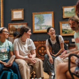 A group of young people smile and talk with each other sitting in a group in a gallery at the Fitzwilliam Museum. Framed landscape paintings hang on the wall behind them.