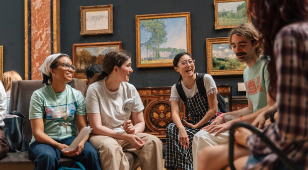 A group of young people smile and talk with each other sitting in a group in a gallery at the Fitzwilliam Museum. Framed landscape paintings hang on the wall behind them.